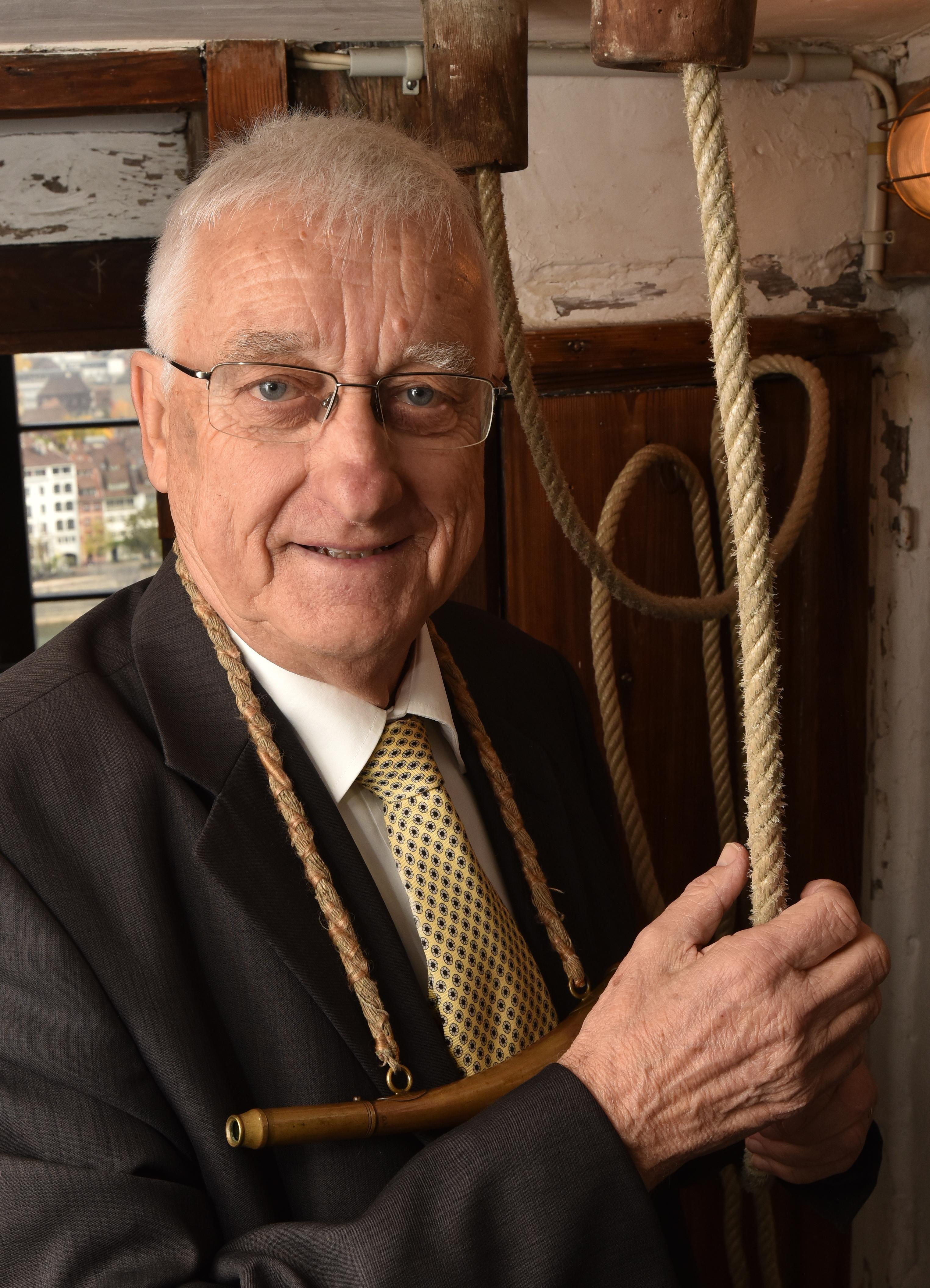 Foto des Messeglöckners Franz Bauer, in schwarzem Anzug mit gelber Kravatte gekleidet, mit dem Horn (Instrument) um den Hals gehängt und dem Seil zur Martinsglocke in den Händen.