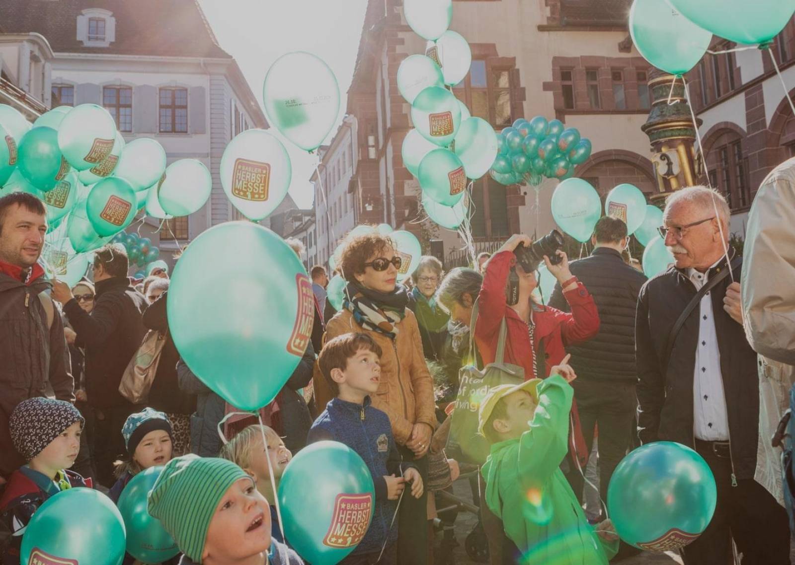 Enfants et adultes regardant la cloche de la foire en train de sonner dans la Martinturm et portant dans leurs mains des ballons turquoise avec le logo rose de la foire d'automne.