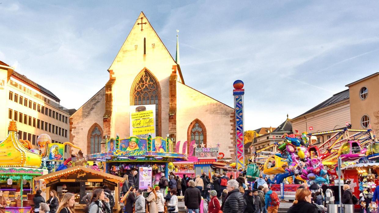 Blick auf das bunte Treiben auf dem Basler Barfüsserplatz, mit farbigen Schiessbuden, Bahnen, Verpflegungsständen und der Barfüsserkirche im Hintergrund.