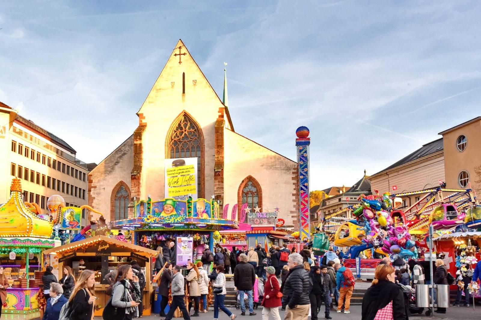 Blick auf das bunte Treiben auf dem Basler Barfüsserplatz, mit farbigen Schiessbuden, Bahnen, Verpflegungsständen und der Barfüsserkirche im Hintergrund.
