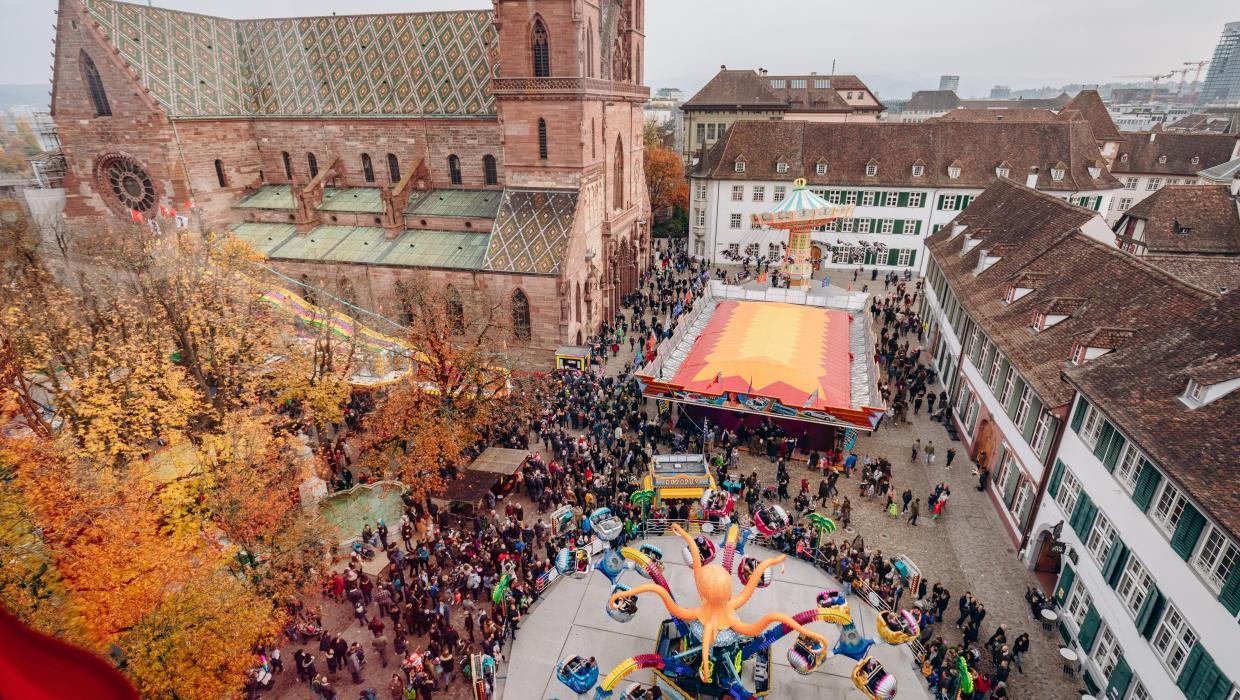Blick von oben auf den Münsterplatz mit seinem herbstlich verfärbten Rosskastanien-Hain und dem roten Basler Münster. Auf dem Platz drehen sich bunte Herbstmesse-Bahnen.