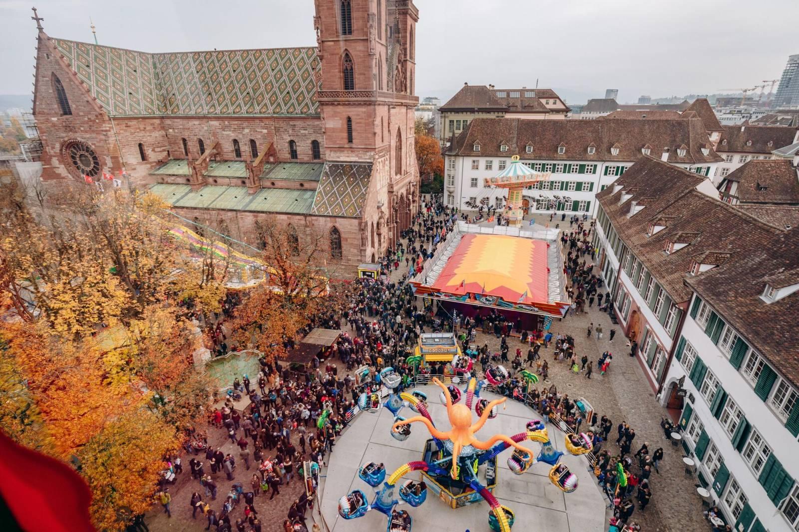 Blick von oben auf den Münsterplatz mit seinem herbstlich verfärbten Rosskastanien-Hain und dem roten Basler Münster. Auf dem Platz drehen sich bunte Herbstmesse-Bahnen.